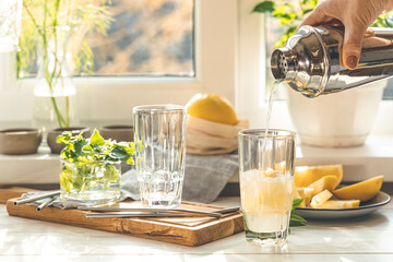 Woman pouring grapefruit gin and tonic cocktail in the glass on the white table near window in the sunny falltime. Selective focus, shallow depth of the field. Alcohol cocktail or detox drink.