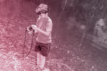 A boy in t-shirt and shorts is standing near the water in nature and cleaning a camera with soap...