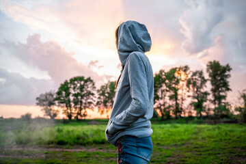 A young woman in a hood stands sideways and looks at the sunset. Forest