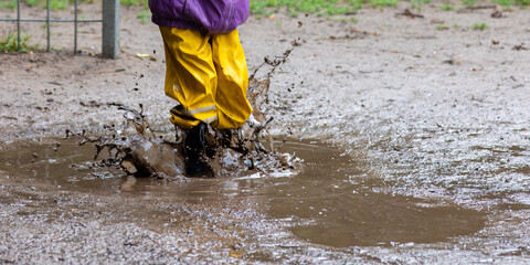 Feet of a Child jumping into a puddle of mud with colorful rubber boots during a rainy and wet...