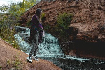 Obraz premium A young woman in a poncho stands near the waterfall and looks at it.