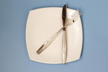 White plate, fork and knife over blue background. Clean plate and cutlery on light background. Top view.