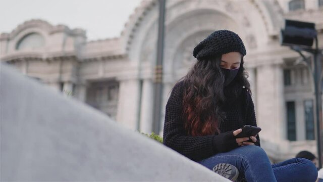 Vista de una adolescente en las calles de la Ciudad de M&eacute;xico con ropa de abrigo y un cubrebocas en una tarde de oto&ntilde;o, covid19, una ni&ntilde;a con su tel&eacute;fono celular jugando o enviando un mensaje