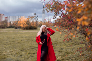 Young beautiful blonde hair woman in red coat walking in autumn park and holding leaves in hand