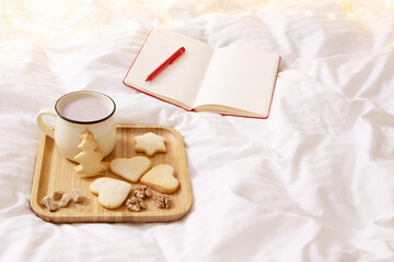 Concept of lazy time at home, holidays. Future planning female theme. Tray with Christmas cookies and cocoa on the bed. Nearby is an open notebook. cotton white blanket, bokeh lights on background
