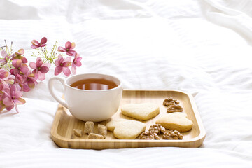 Fototapeta na wymiar Good morning, wonderful day concept. Breakfast in bed: cup of fragrant tea with homemade delicious cookies, brown sugar on wooden tray. Pink flowers, open book a white blanket on the background.