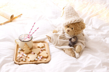 Fototapeta na wymiar Christmas, milk for Santa concept. Winter food in bed: cup of tea, jar of milk, homemade cookies on wooden tray. Toy bear in knitted hat, open book. White blanket, garland lights in the background.