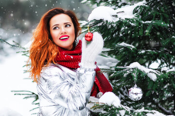 Outdoors lifestyle portrait of stunning redhair young woman. Smiling and decorating of christmas fir-tree. Wearing stylish scarf, silver down jacket. Christmas mood. It's snowing. Happy new year