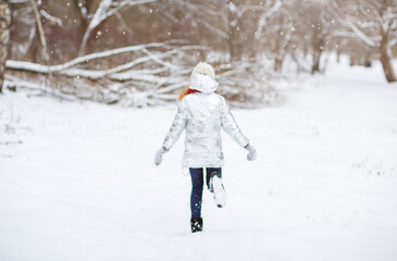 Winter lifestyle portrait of cheerful pretty girl throwing snow in the air. Smiling and having fun in the snow park. Snowflakes falling down. Wearing stylish knitted hat, silver down jacket, mittens