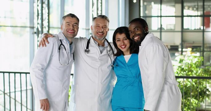 Portrait of happy mixed-races males and female doctors standing in clinic, looking at camera and laughing in hugs. Cheerful multi ethnic men and woman physicians embracing and smiling in hospital.