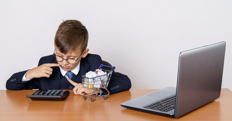A young businessman is counting on a calculator. There is a computer and a mini shopping trolley on the table.