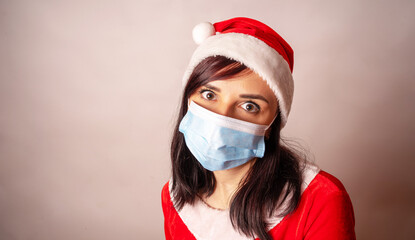 Fototapeta na wymiar Young woman in medical mask and Santa Claus suit on grey background. Close up of female in Christmas hat and protective mask.Concept of safe Christmas celebration during coronavirus pandemic.