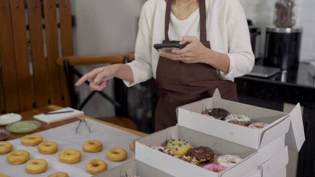 beautiful asian woman smiles wearing an apron making donuts and receiving food order through mobile phone