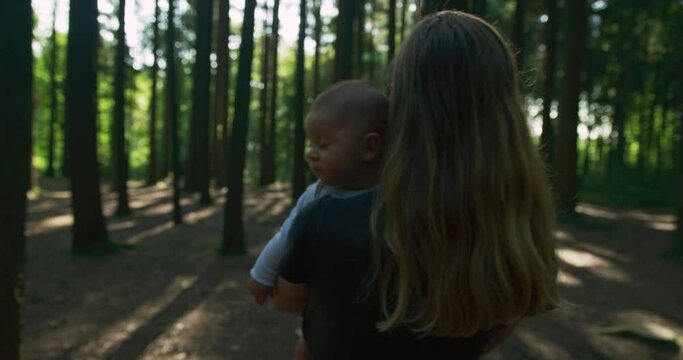A young mother is walking and carrying her baby through a forest on a sunny summer day