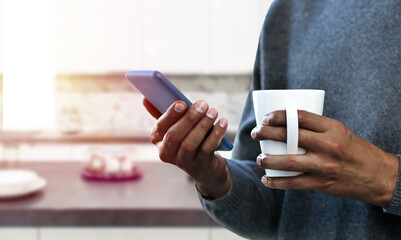 Man hands holding a coffee mug with phone inside of a kitchen