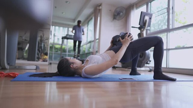 Young Asian female doing hip raise bridging exercise for strong and tight core lower back muscle, holding weight lying down on exercise mat, indoor fitness club public gym, fit and firm body