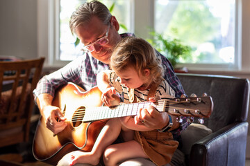 Grandpa and granddaughter playing guitar in the living room in natural sunlight - 387493539