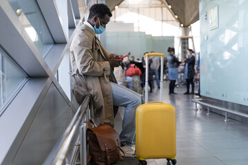 Afro-American traveler man in trench coat wear face protective mask, sitting in airport terminal or...