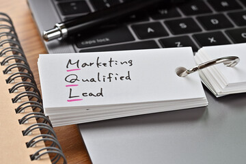 The words "MQL" written in a word book with laptop PC and a pen. It is an acronym for "Marketing Qualified Lead".