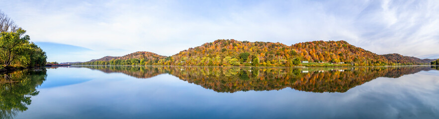 Hills covered in colorful fall foliage are reflected on the still water of the beautiful Ohio River. Monroe County, Ohio is viewed from the river bank at Paden City, West Virginia. - obrazy, fototapety, plakaty