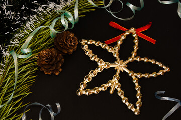 Golden christmas tree star with red bow on black background near green pine branch and cones with blue curly ribbon and tinsel. Christmas preparations concept. Christmas tree decorating