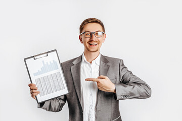 Young handsome brunet man in glasses in suit smiles and points at business with documents and graphs on white isolated background