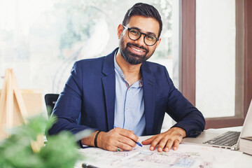Handsome bearded Indian architect working from home on a building blueprint and laptop
