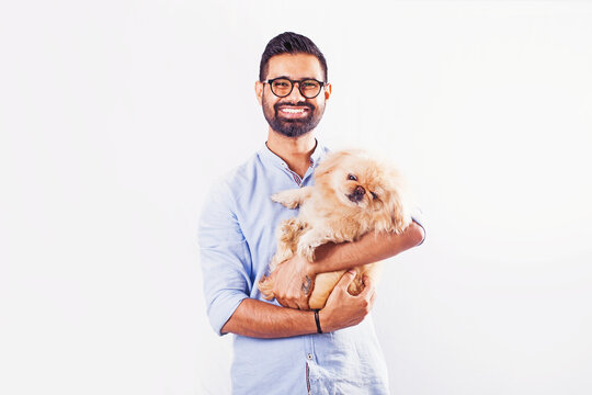 Young handsome Indian man holding a pekingese puppy over white studio background