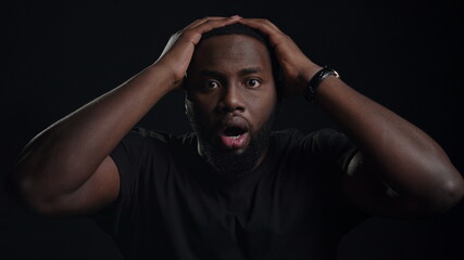 Stressed afro guy posing on black background. Guy holding hands on head indoors