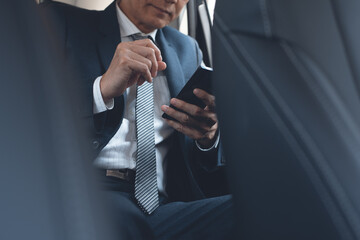 Asian businessman in black suit using mobile phone inside a car on backseat