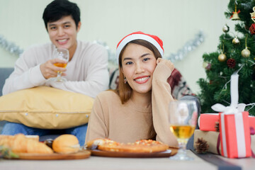 Obraz na płótnie Canvas close up young asian woman sitting on dining table with man sit on sofa in living room with decorative Christmas pine tree for celebration party at home concept