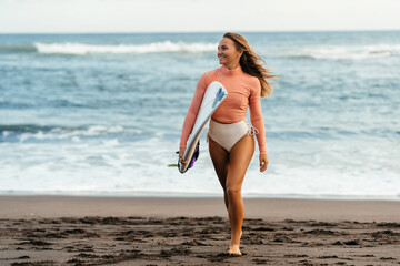 Young attractive surfer woman with white board at sunset on the ocean. Bali Indonesia. Summer time, sports, travel content.