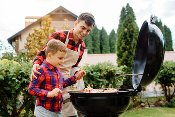Young father and his son are cooking barbecue on an outdoor grill in their home backyard and having...