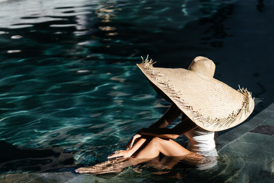 Woman in big straw hat relaxing at pool. Wellness and relaxation concept for summer luxury vacations.