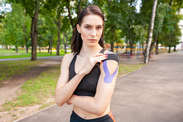 Young stunning muscular woman wearing black sports outfit. Fit brunette gorgeous female athlete putting colorful kinesiology tapes on shoulders. Concept of sports treatment, rehabilitation.