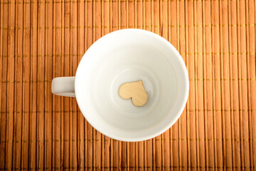 Fototapeta na wymiar Top view of one white cup with a small wooden heart on a table, in warm light, brown monochrome indoor background photographed with selective focus 