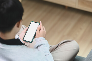 A young female is relaxing on sofa while holding smartphone mock up blank white screen at home.