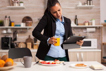 Multitasking woman talking on the phone, holding tablet and drinking a juice. Concentrated business woman in the morning multitasking in the kitchen before going to the office, stressful way of life
