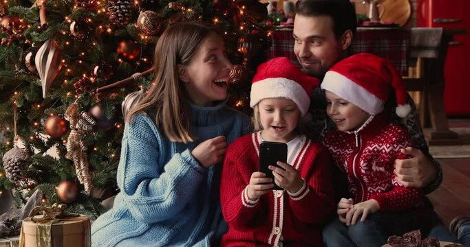 Happy young couple and small kids sit on floor near glowing bright decorated Christmas Tree using smartphone take photography capture moment New Year celebration, family having fun on xmas eve concept