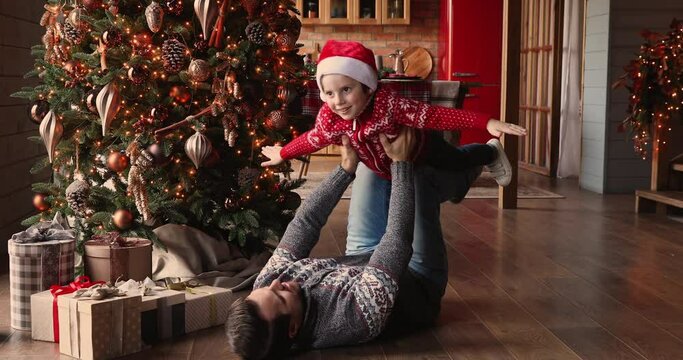 Father lying on warm wooden floor in living room decorated with lights glowing Christmas tree lift up small son wear Santa hat and sweater, family having fun play at home enjoy happy holidays concept