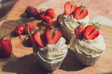 Appetizing cupcakes with white cream decorated with fresh strawberries lit by the rays of the sun