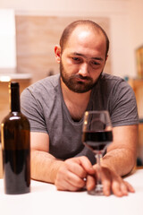 Man sits depressed and alone with a glass of red wine at kitchen table. Unhappy person disease and anxiety feeling exhausted with having alcoholism problems.