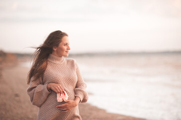 Fototapeta na wymiar Pregnant woman wearing knitted comfy sweater holding baby shoes walking at sea beach outdoors close up. Motherhood. Maternity. Healthy lifestyle. Healthcare.