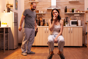Desparate wife smoking after being beaten by alcoholic husband in kitchen. Violent aggressive man...