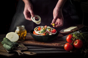 Cooking process of Greek Salad for a restaurant menu. Food composition of a Mediterranean cuisine in rustic style. Chef put the onion and olives in a black bowl and mixed of all vegetables in a salad.