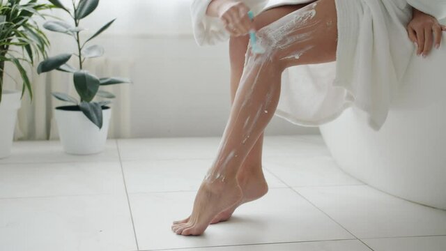 Close-up of female legs covered with shaving foam and hand removing hair with razor caring for skin and beauty in bathroom. People and lifestyle concept.