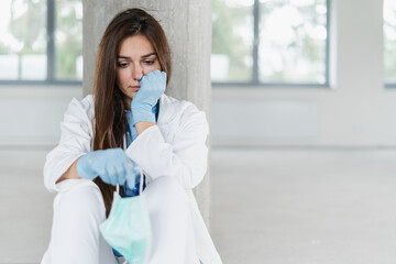 Sad and hopeless young female doctor holding protective mask for protecting from virus desease sitting on floor at hospital.