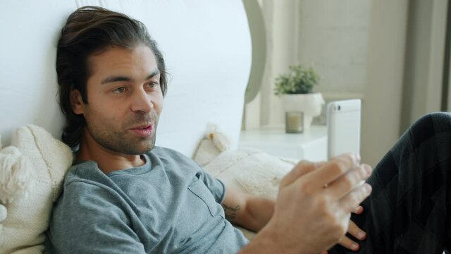 Attractive mixed race man is making online video smart phone call talking and waving hand in bed at home. Communication and people concept.