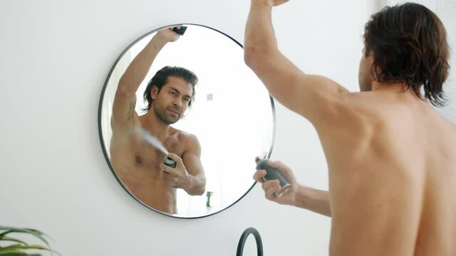 Handsome young man is using deodorant spray in bathroom applying on armpits looking at mirror at home. Hygiene and modern people concept.