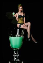 Pretty sly green fairy sitting on glass of absinthe. Portrait of sexy smiling young girl on black...